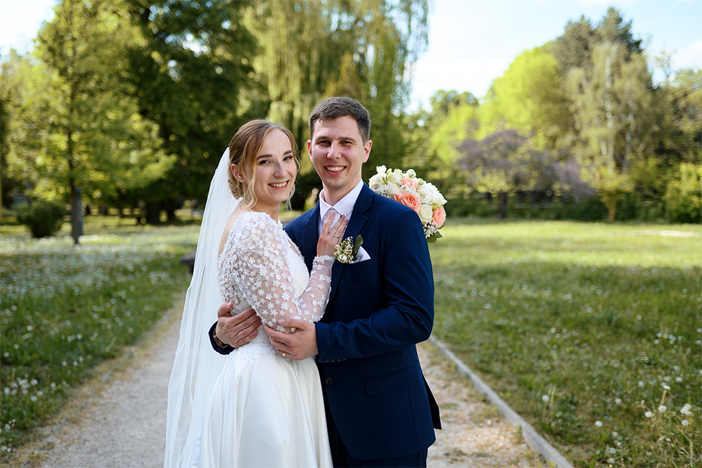Usmievavý ženích a nevesta v objatí na chodníku uprostred parkovej zelene.