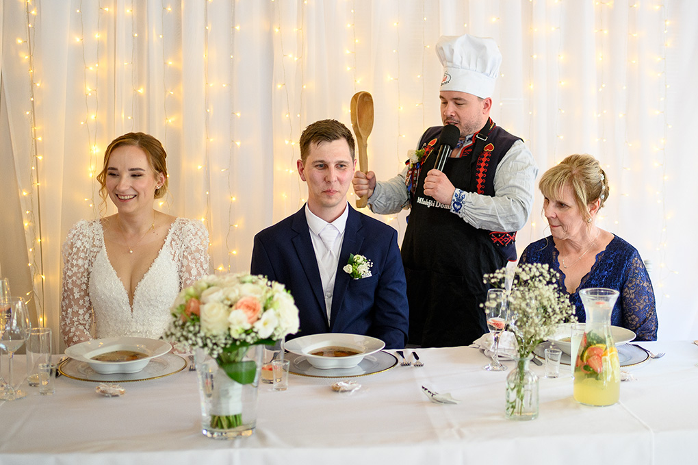 Starejší v kuchárskej čiapke a zástere drží veľkú drevenú lyžicu nad ženíchom.