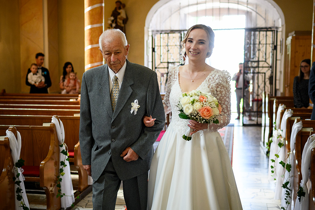 Nevesta a svadobný otec kráčajú uličkou medzi lavicami k oltáru.