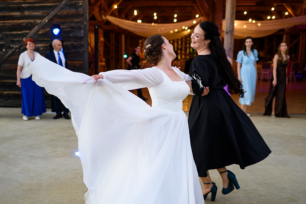 Nevesta roztáča sukňu počas tanca so svadobnou hostkou pred otvorenou stodolou.