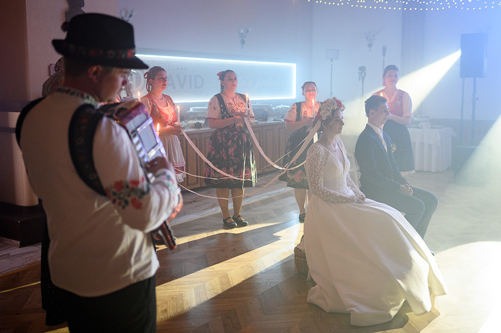 Pohľad na usadený pár cez harmonikára v popredí počas vystúpenia folklórneho súboru.