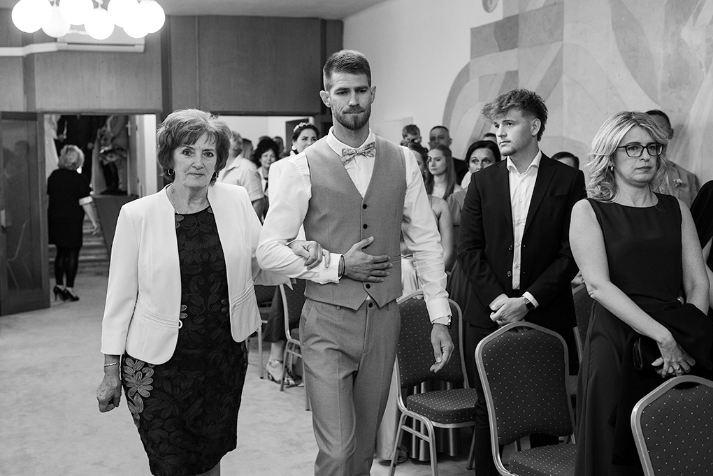 Ženích v obleku s vestou kráča uličkou zavesený do svojej mamy pred začiatkom obradu.