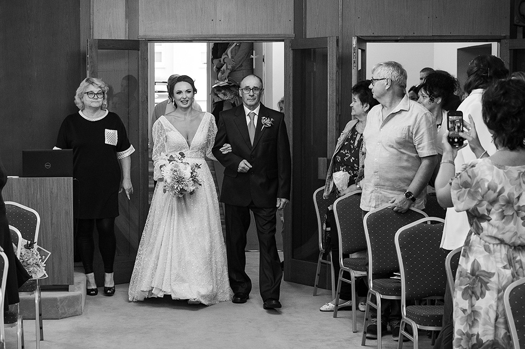 Nevesta vchádza do sobášnej siene v sprievode svojho otca, hostia sa prizerajú.