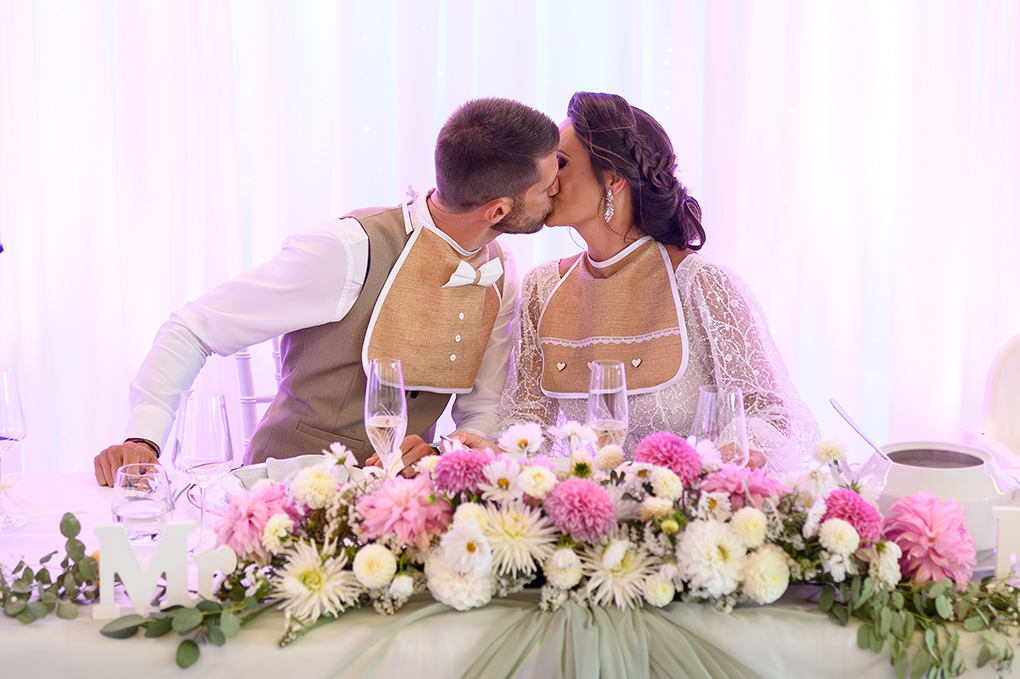 Romantický novomanželský bozk pri hlavnom stole s bohatou kvetinovou výzdobou.