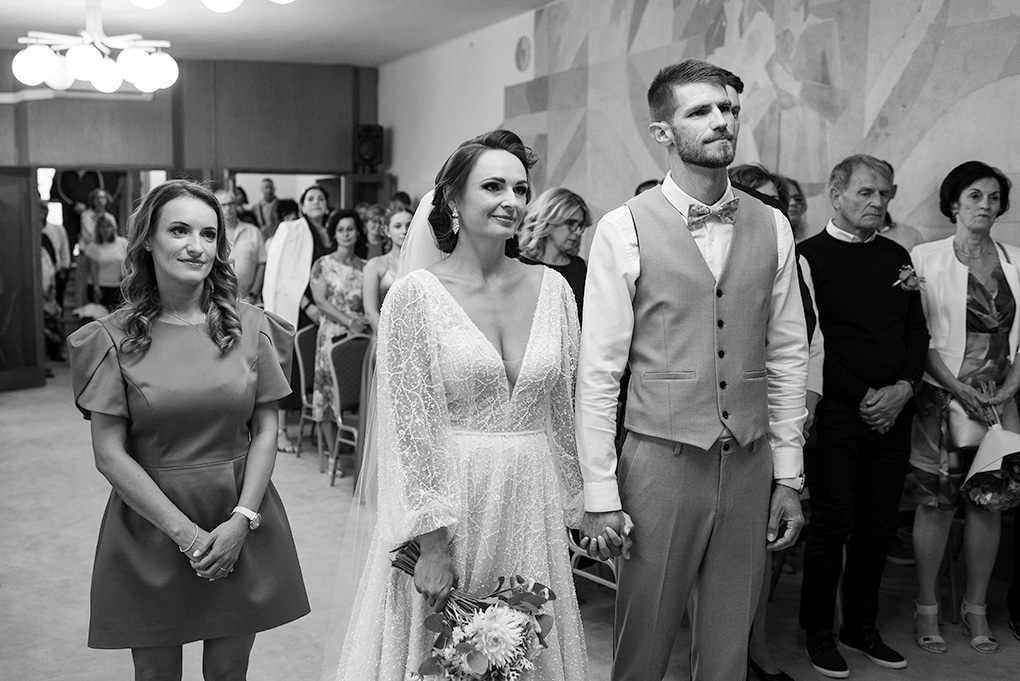 Mladomanželia sa držia za ruky a počúvajú príhovor oddávajúceho.