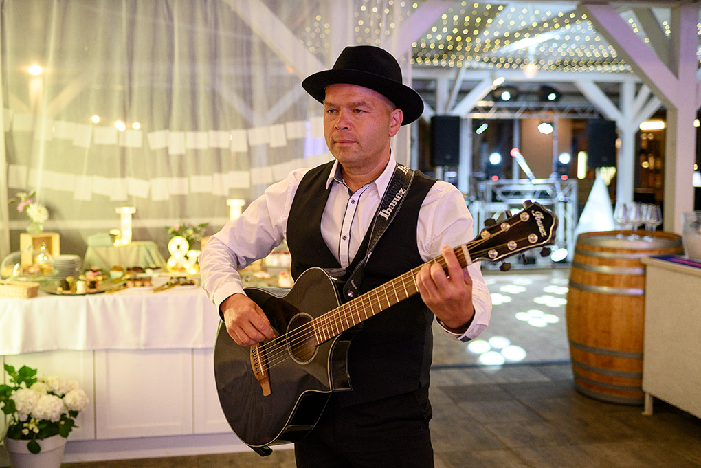 Svadobný hudobník v klobúku hrá na gitare počas večerného programu.